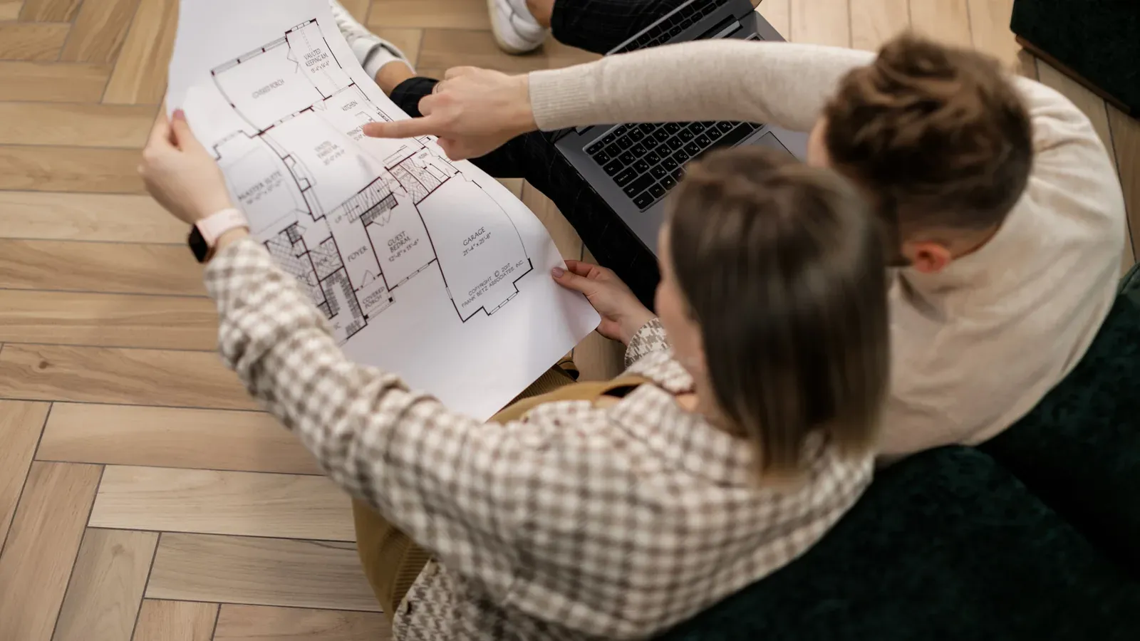 Ein Paar sitzt mit Bauplan und Laptop auf dem Boden und plant den Hausbau